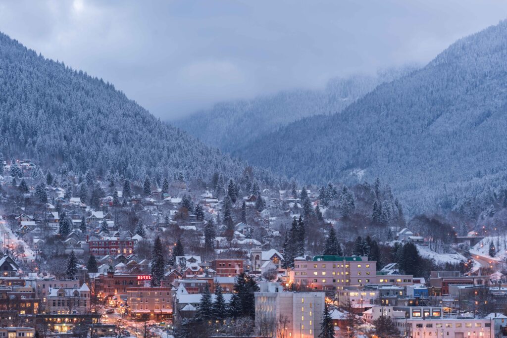 A snowy townscape at dusk, with snowy tree covered mountains surrounding valley town