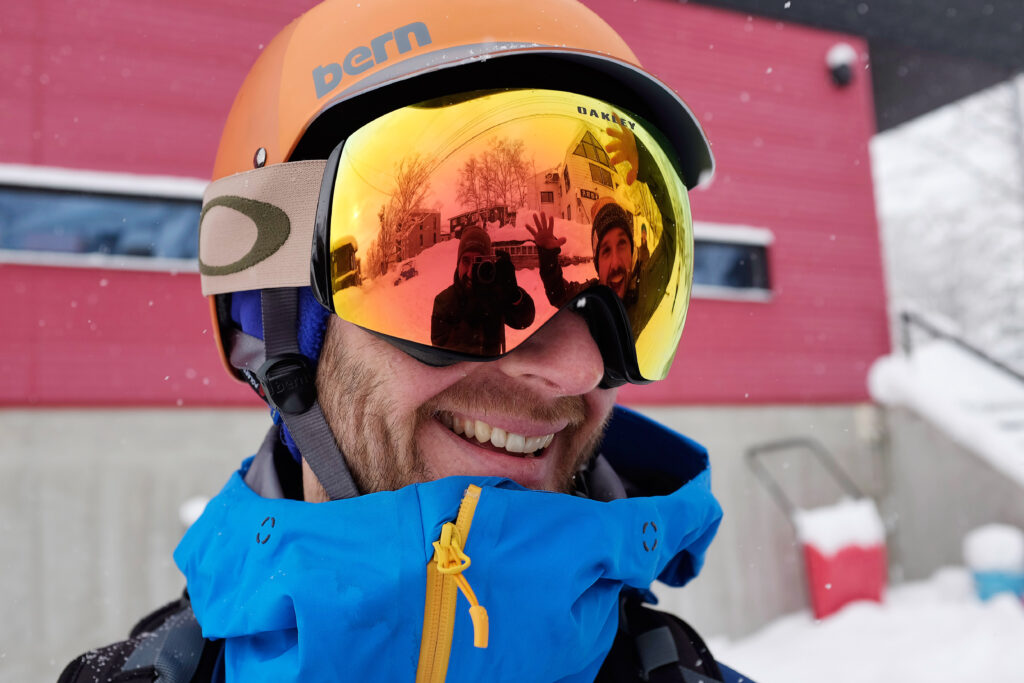 man wearing orange helmet and orange-yellow lense goggles pictured up close, smiling, as two others are photographed in reflection, one taking photo, second pulling a face with arms raised. 