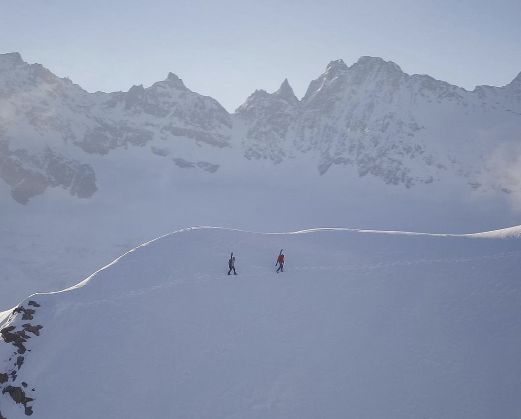 image taken by drone high in mountains with two ski tourers as tiny figures traverse a snowy ridge in the shade, the peaks of tall mountains make the backdrop ridge behind
