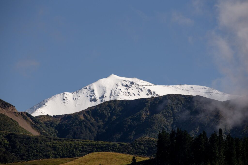snowy mt hutt ski resort covered in snow is set back against the positively summery hill in front, green with summer foliage