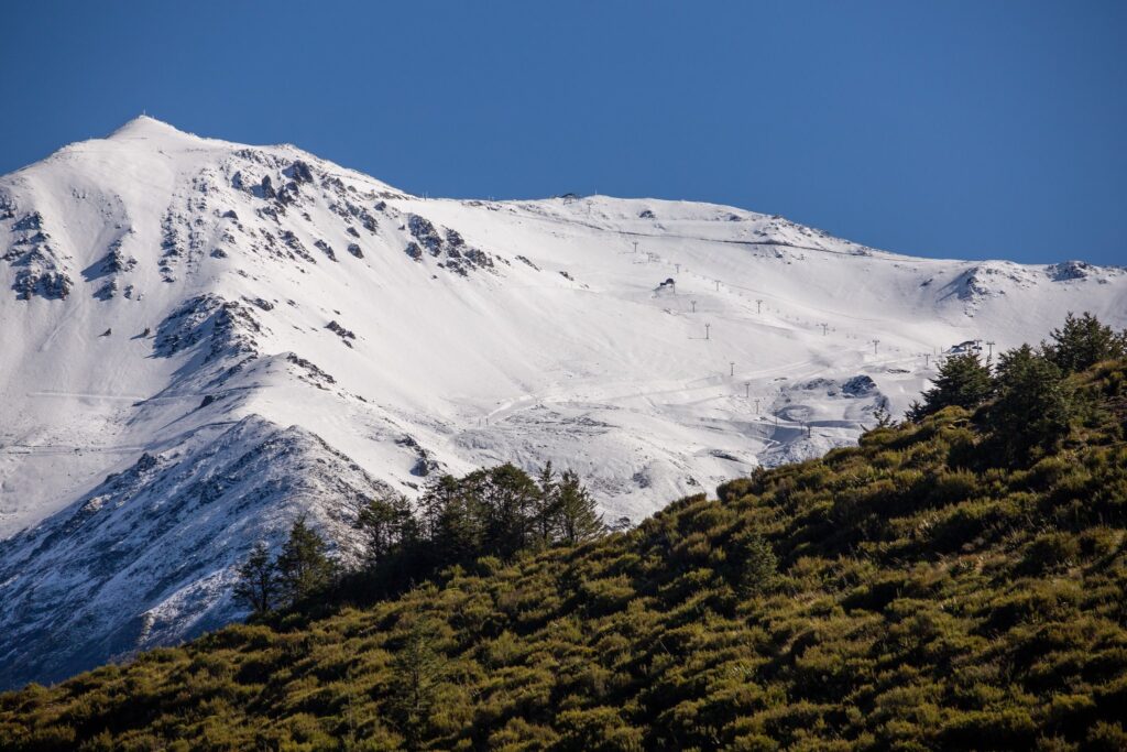 snowy mt hutt ski resort covered in snow is set back against the positively summery hill in front, green in colour from summer foliage