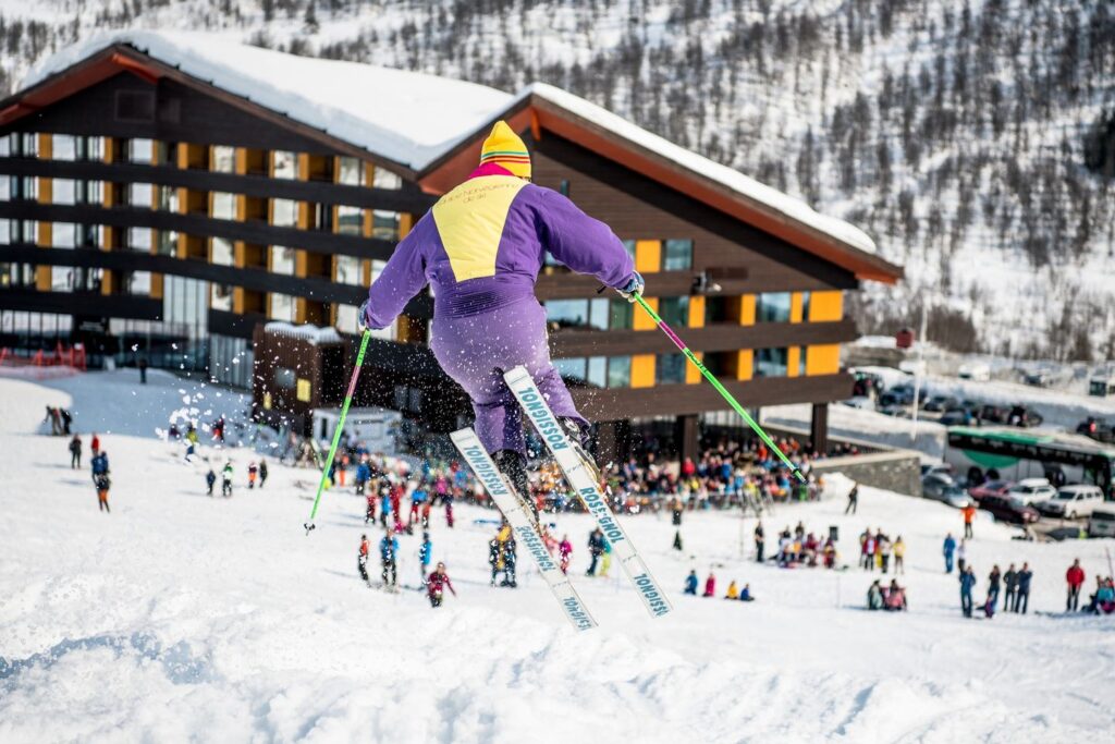 skier hits a bump and takes a little air, wearing a retro onesie in front of a base-building with people scattered below at end of piste
