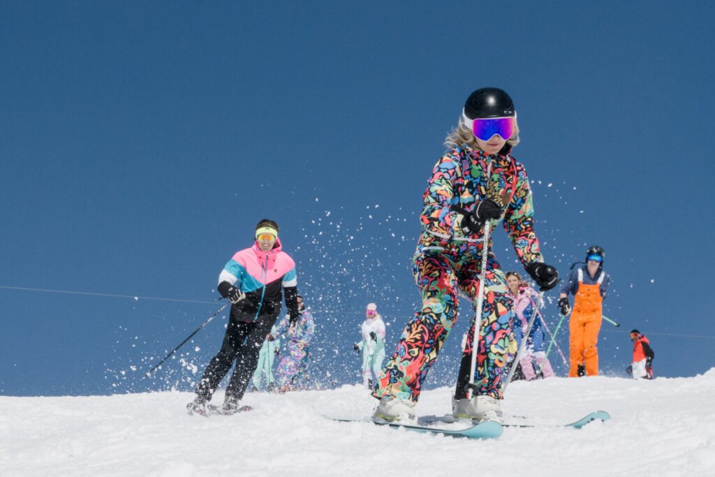 posse of skiers head down a easy slope in formation around a camera placed on piste all wearing loud, patterned and fun ski gear