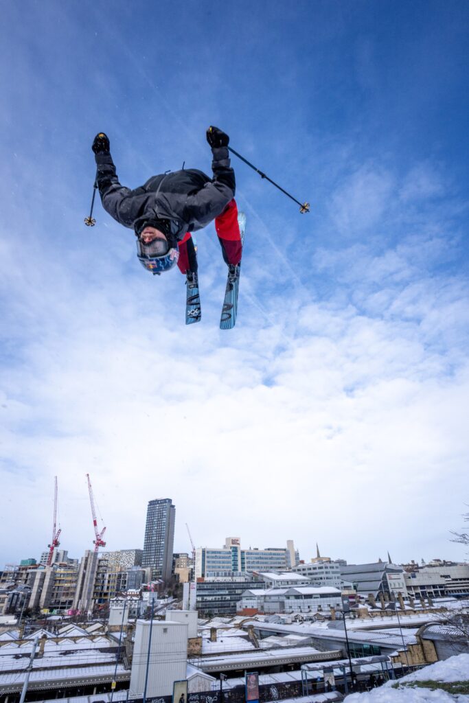 skier backflips off a kicker in UK's Sheffield with city scape behind.