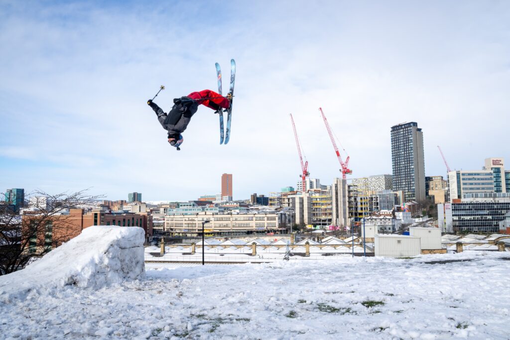 skier backflips off a kicker in UK city Sheffield with city scape behind.