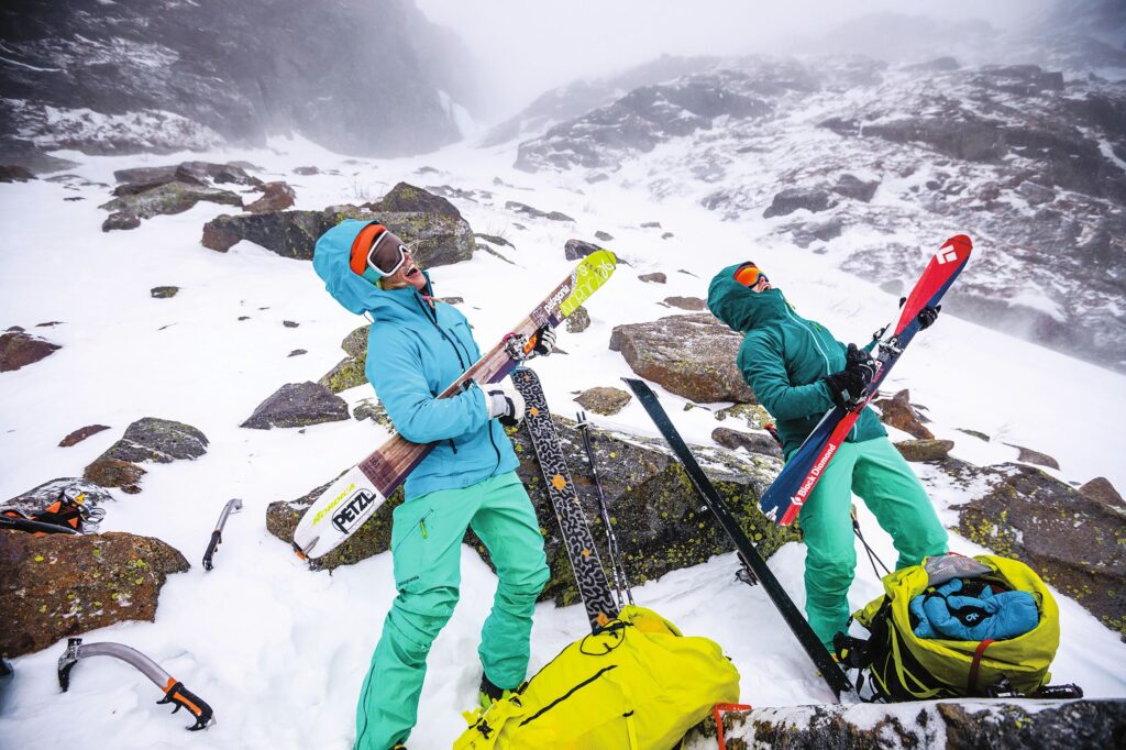 two skiers in bright blue and green play their skis like air guitars on a rocky-snowy face