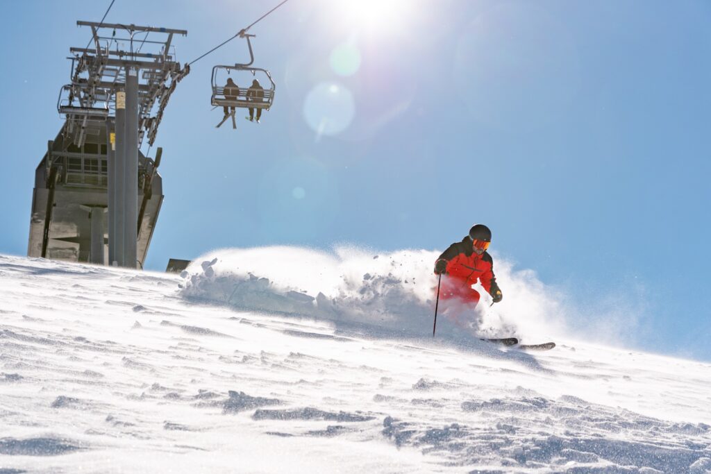 a skier in red and black gear skis in resort under a chairlift with a light powder spray behind him