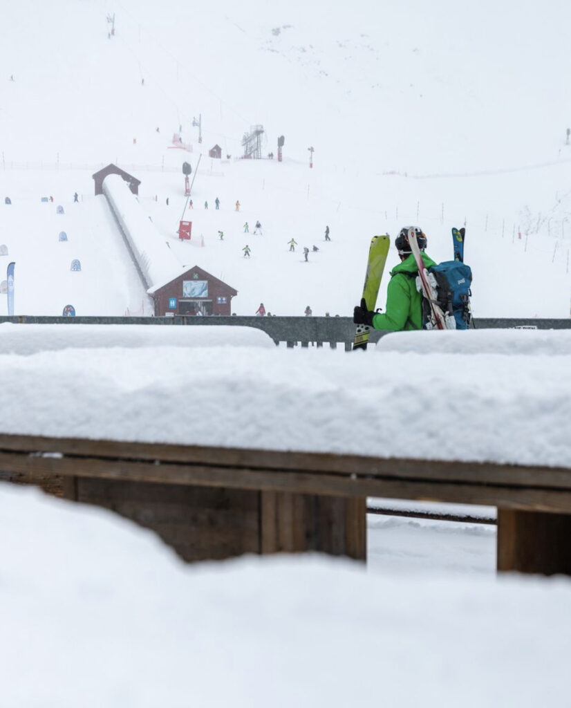 skier in green carries yellow skis towards ski lift, but the foreground of image is a barrier, bannister covered with 5cm or so of snow, to show recent snowfall