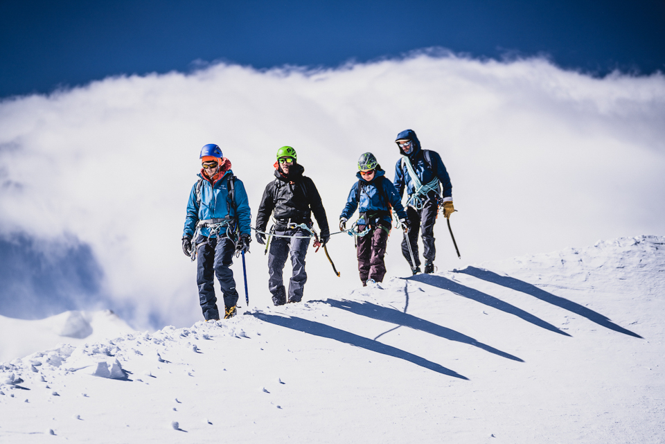 four mountain alpinists walk along a snowy ridge, roped up, carrying ice axes and wearing helmets and sun glasses. Just sky and cloud behind them, so they must be high