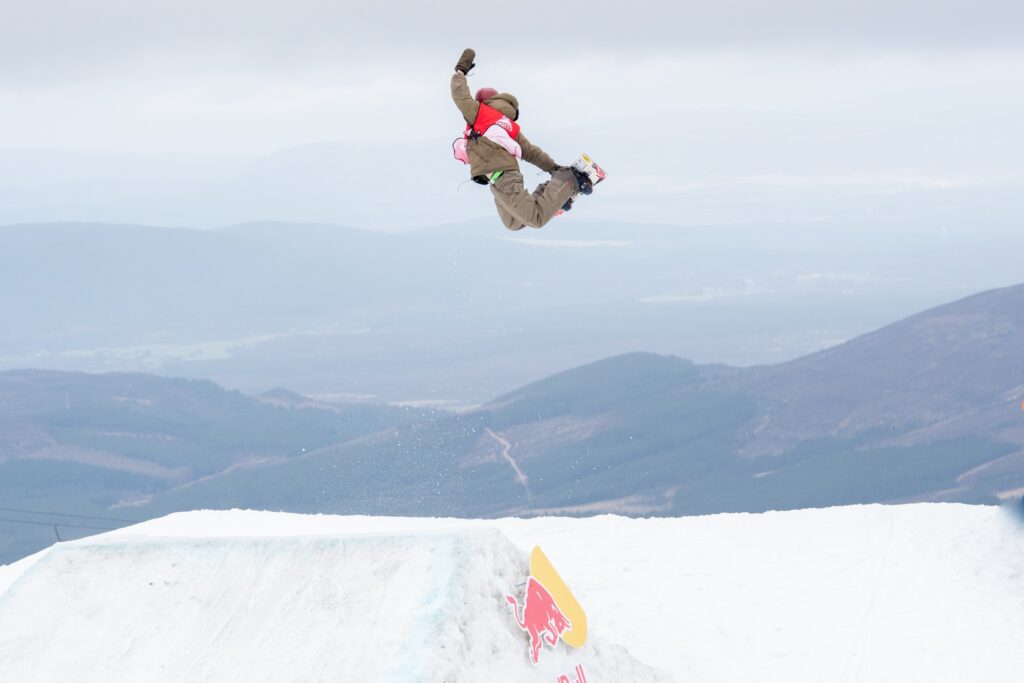 red bib BRITS competitor mid-jump grabbing heel edge of snowboard with a non snowy landscape in distance
