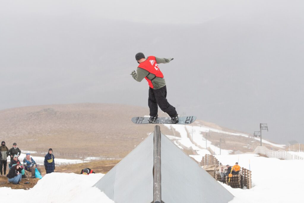 red bib BRITS competitor mid-rail grind with a less snowy hill in distance.