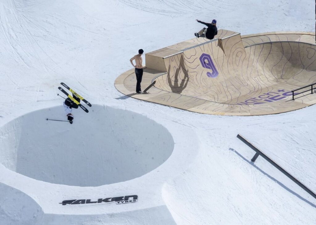a skier upside down over a mad jump in The Nines competition, with a snowboarder behind on different kicker/skate bowl feature