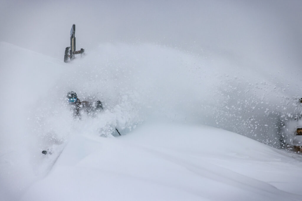 A skier isn't really visible through the snow they're kicking up from a turn. Powder sprays downhill and perhaps a sign is visible through the whiteout, snow-spray