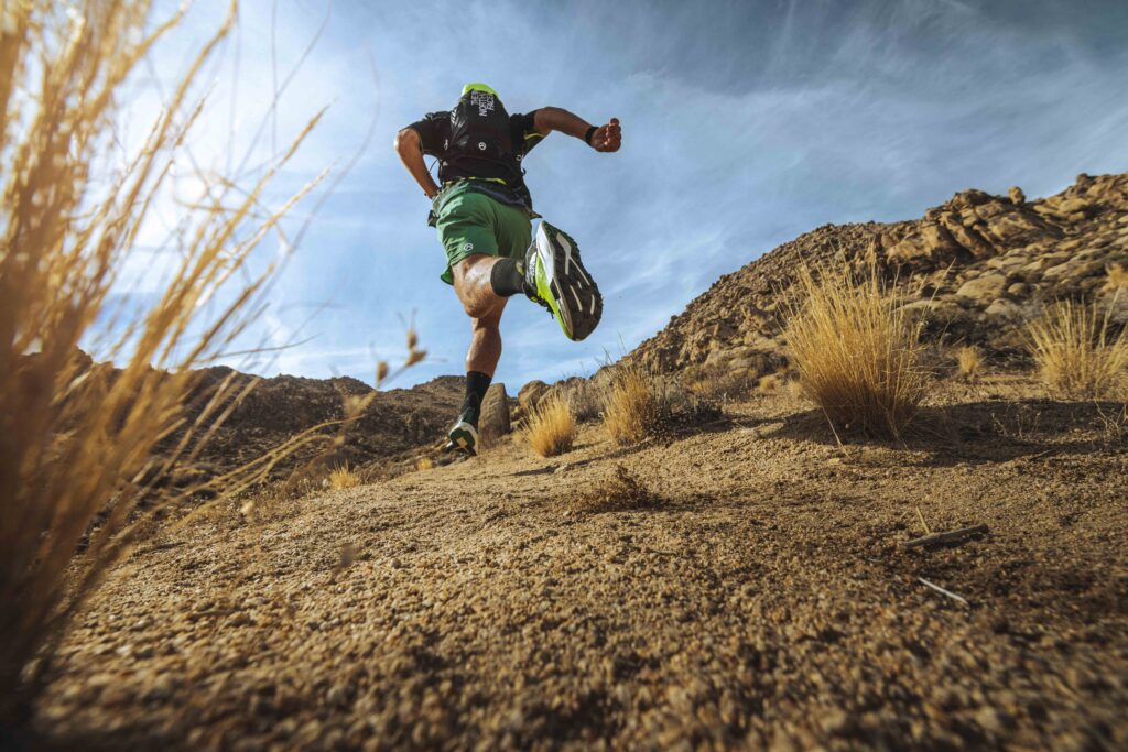 A trail runner is mid-stride on sandy, gravelly terrain with dry grass, uphill running. He's wearing green and black The North Face running gear, and is silhouetted against a blue sky
