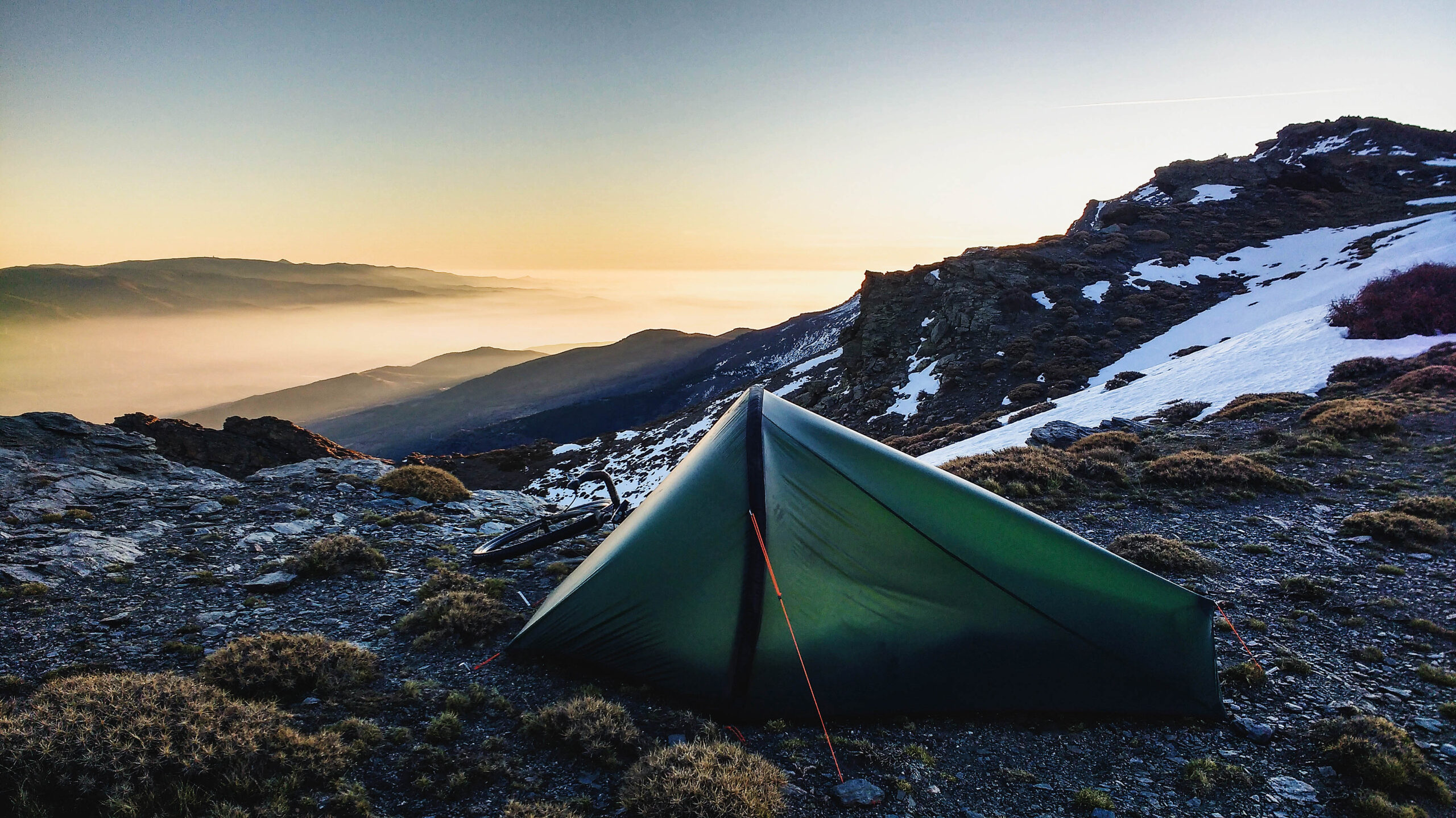 A green tent sits erected at the top of a mountain, with snow in the background in patches, as well as bushes. It looks like dusk/dawn as the sun casts an orange glow over the mountains in the distance