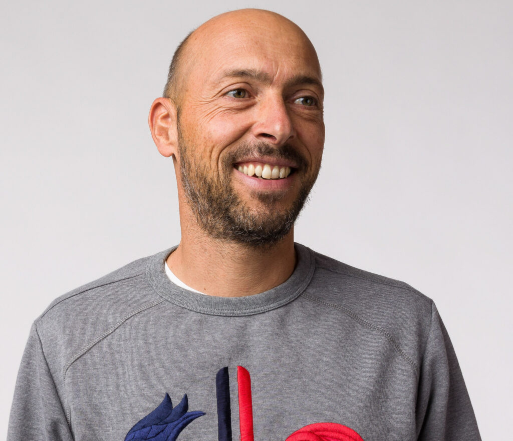 A man smiles to the side of the camera wearing a grey jumper with the Rossignol brand symbol on, in a profile shot