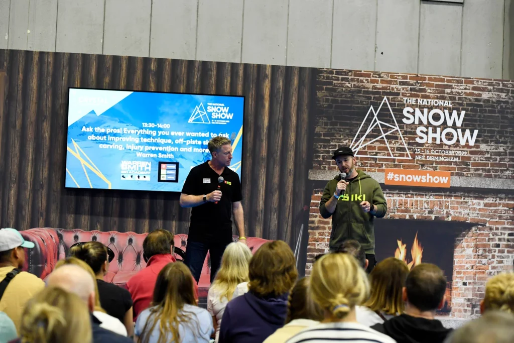 two speakers on stage at the National Snow Show, photo taken from behind with the backs of audience