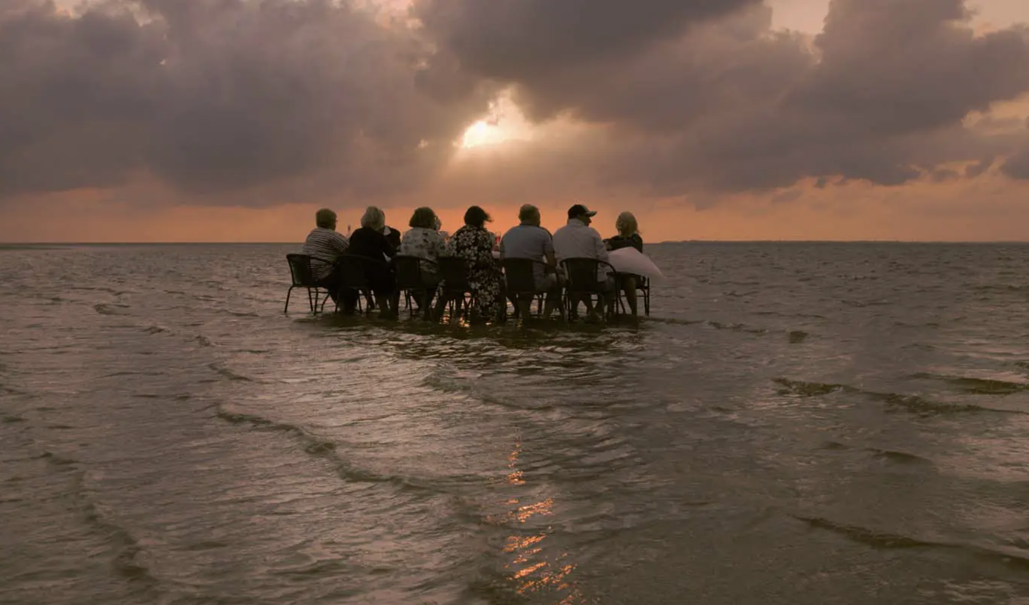 a row of chairs with people sat on them are lined up in the shallow water of a sea with the changing tide, during an orange-glowing sunset sky