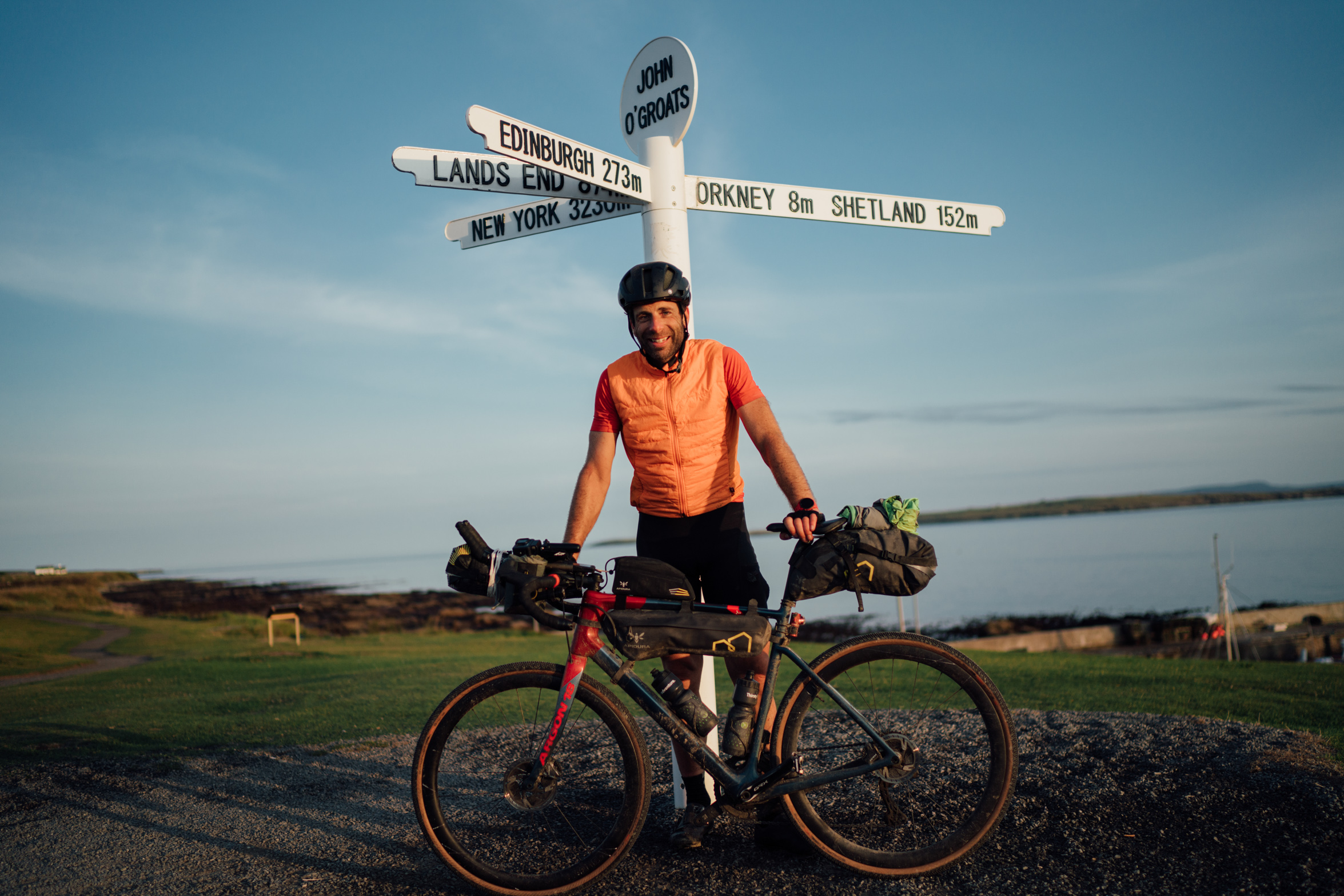 A man in orange stands with bike under a road sigh in John O Groats, with the distance to various other destinations on the multi-pronged sign post. The sea sits behind him, blurred