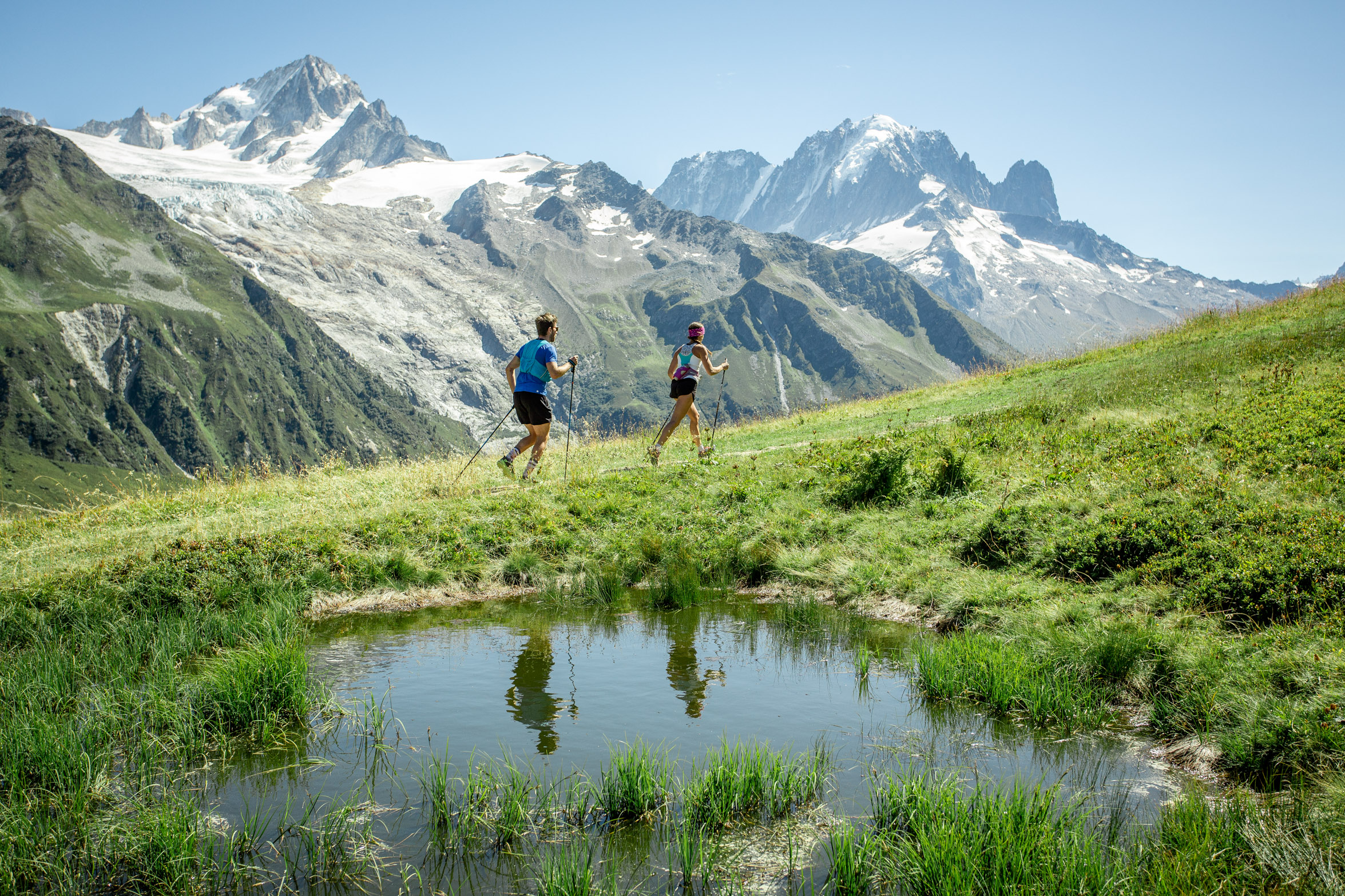 two mountain runners/speed hikers with sticks make a quick ascent, their reflection in a small mountain lake in the foreground, the snowy peaks of high mountains behind then, as they run along a grassy trail