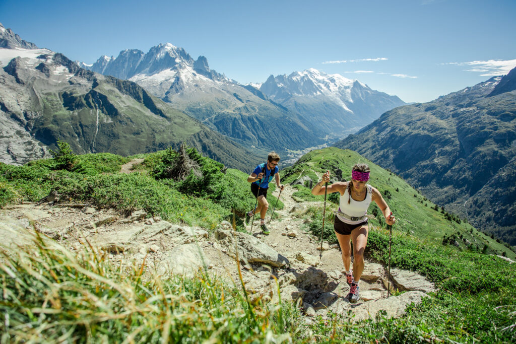 two mountain runners/speed hikers with sticks make a quick ascent, the snowy peaks of high mountains behind then, as they run along a dry dirt trail with green grass on the side