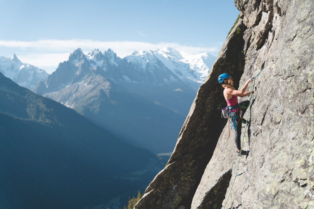 A climber is mid-way up a sheer rock race, looking up. High snowy capped mountains are in the distance beyond rock wall, showing how high this roped-up climber must be