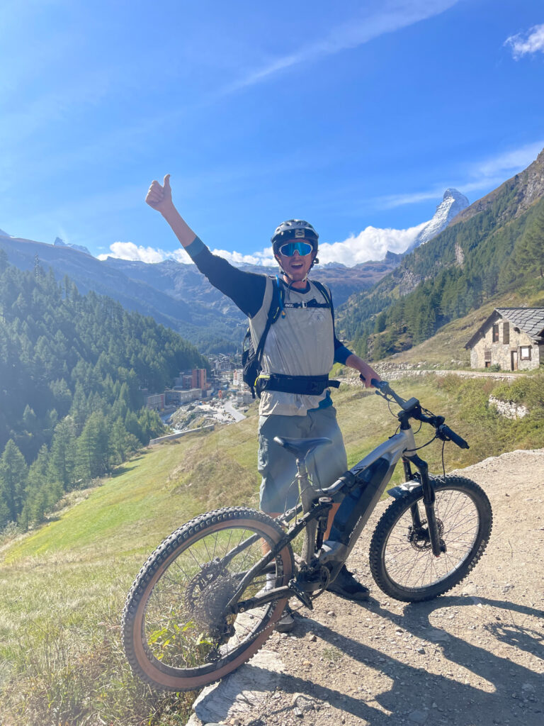 biker gives a thumbs up as he stands behind bike, the snowy matterhorn mountain just visible in the distance, with green slopes and a stone house closer by
