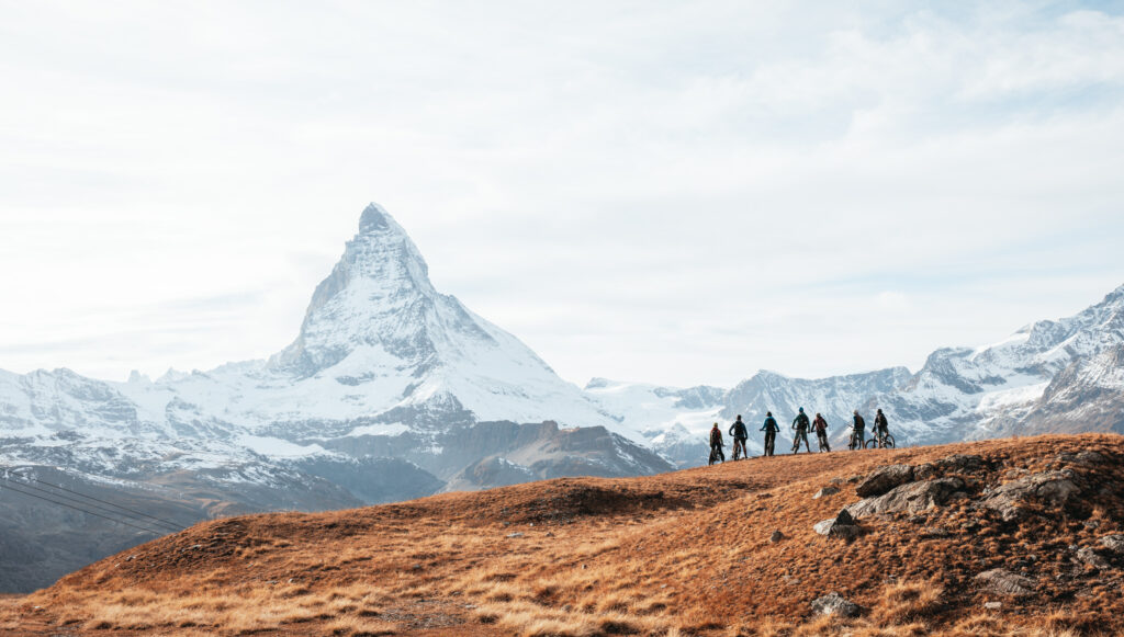 Seven biker silhouettes stand on a ridge of golden grass, looking across at the snowy ridge and peak of the Matterhorn