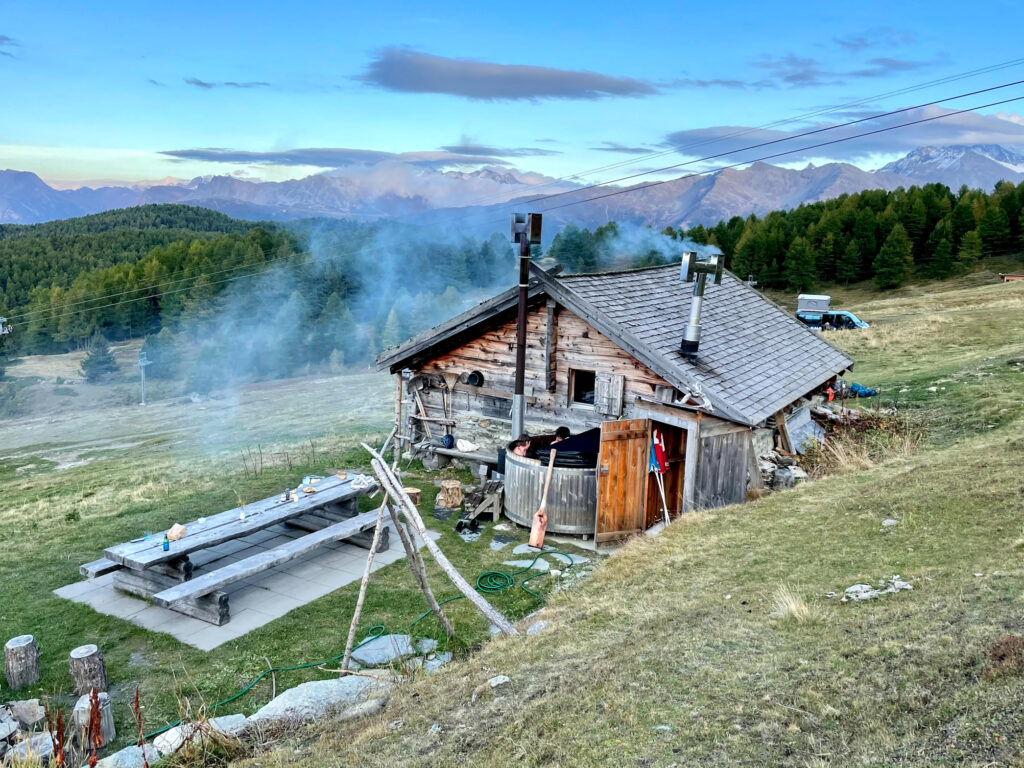 a tiny mountain hit stands in a grassy field in the mountains, looking very quaint and rustic, steam rising out of chimneys