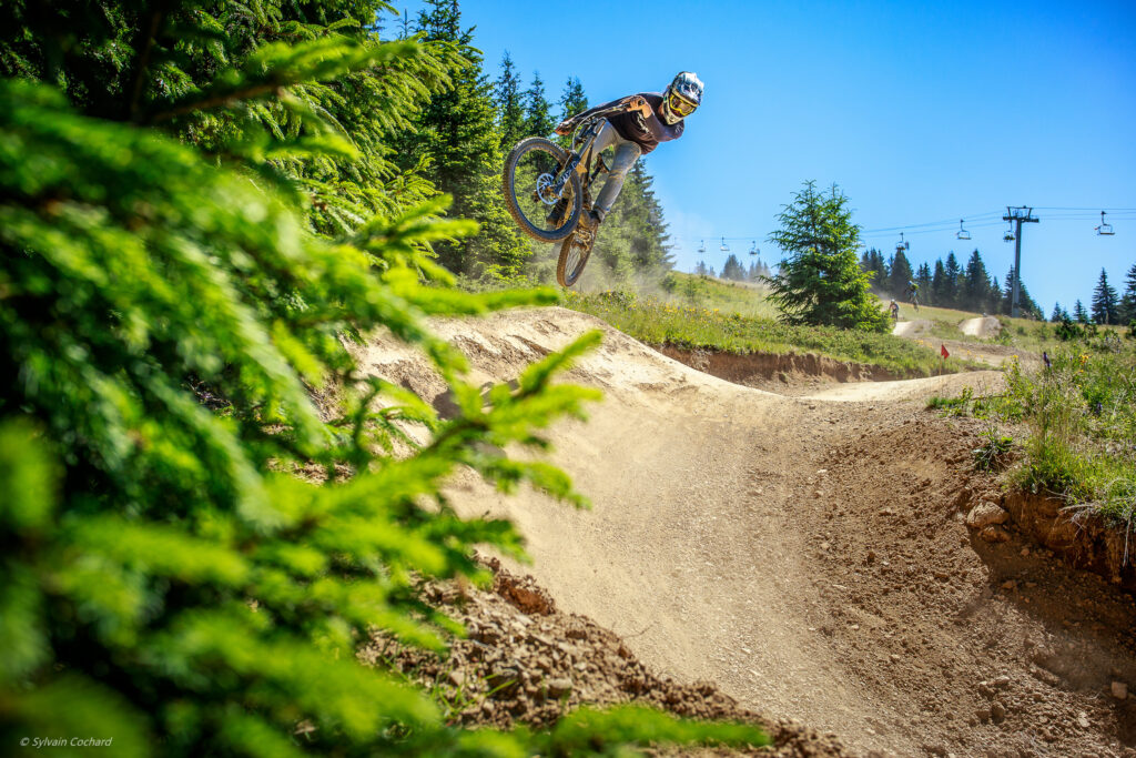 A mountain biker takes air off a berm on a trail, the camera hidden half behind leaves and down at ground level