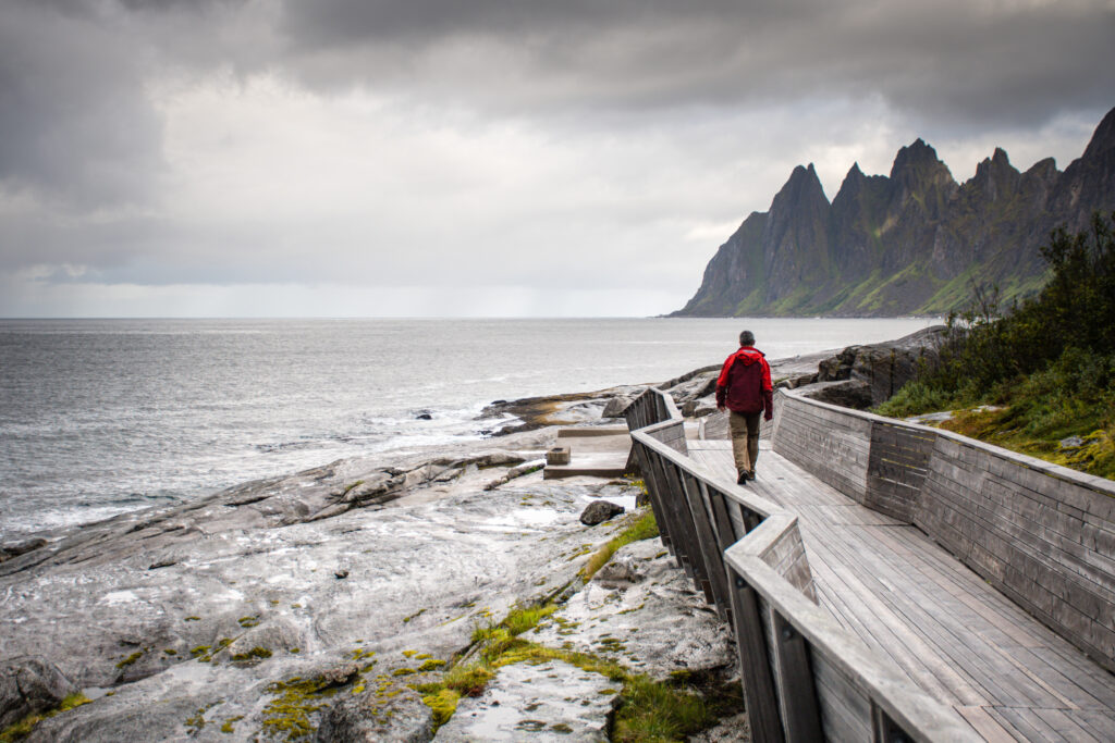 A man in red walks along a wooden walkway along the coastline, mountains and jagged peaks ahead of him