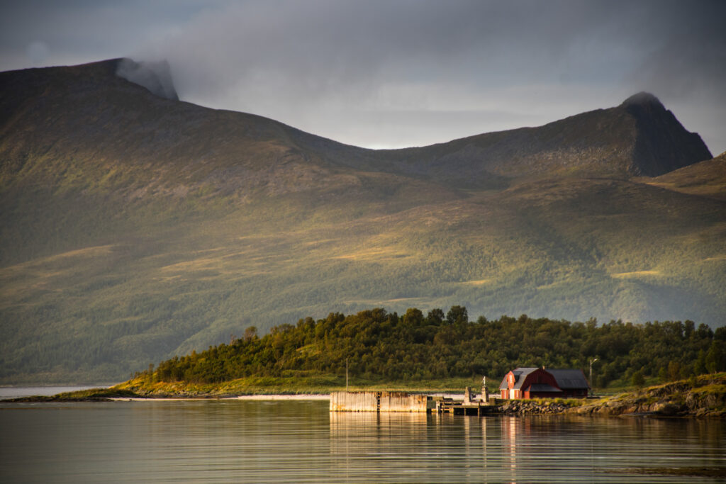 A warm light is cast over a lone red barn sitting on the coastline, green-covered mountains behind it