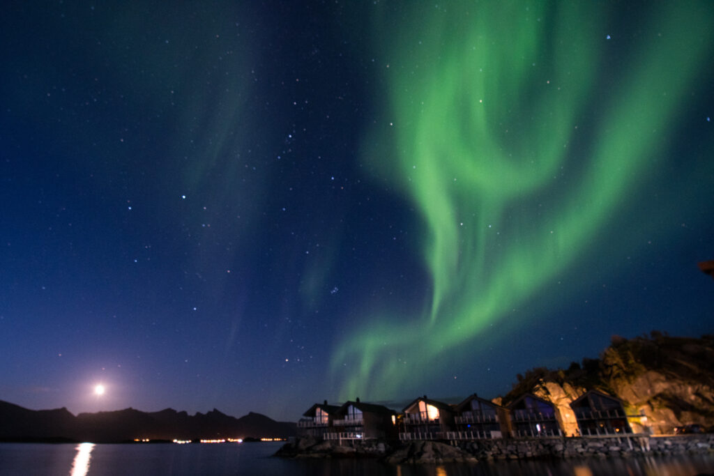 Green swirls of Northern Lights mark the sky, blue in colour over sea and small human settlements lit up with electric lights. The moon shines low over the hill tops