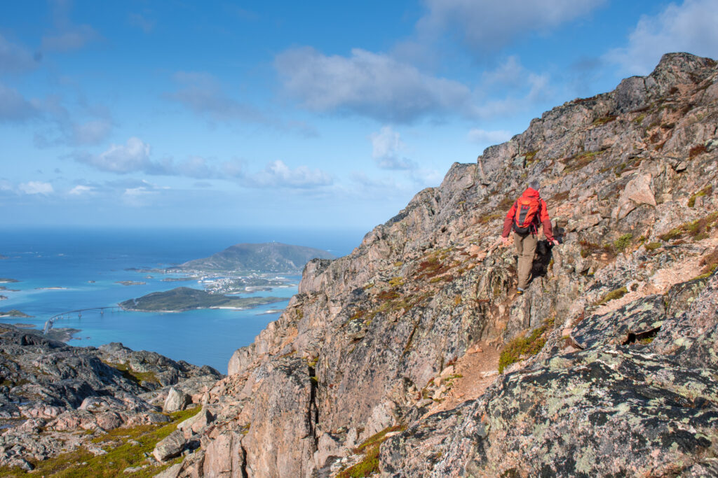 A climber in red climbs a rocky mountain top, sea and islands visible far below