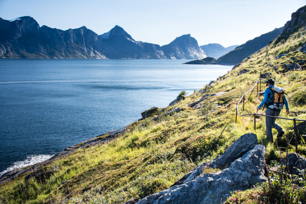 A walker on a pathway next to blue sea and mountains on the other side (landscape is fjord)