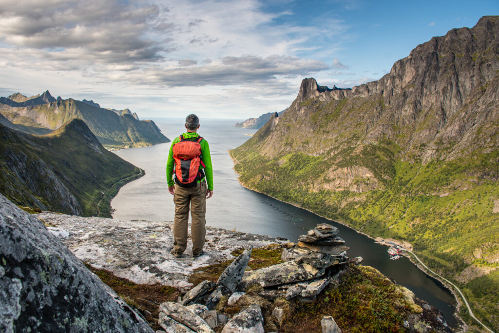 A man stands on a flat rock looking over the sea on a mountain top. The landscape is fjords