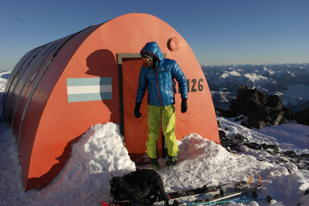A man in yellow and orange stands outside an orange metal shelter with the Argentina flag painted on, at the top of a snowy mountain