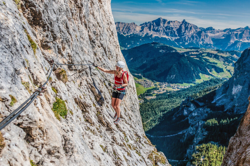 A woman very high on a rock face, travels through a via ferrata course, the valley hundreds of metres below