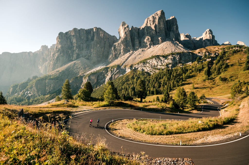 Winding alpine hairpin turn type road, with two cyclists heading downhill, towards the huge monolith rock that characterises the Dolomites