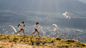 two trail runners run uphill along a grassy mountain ridge, high in the mountains, the valley visible far below
