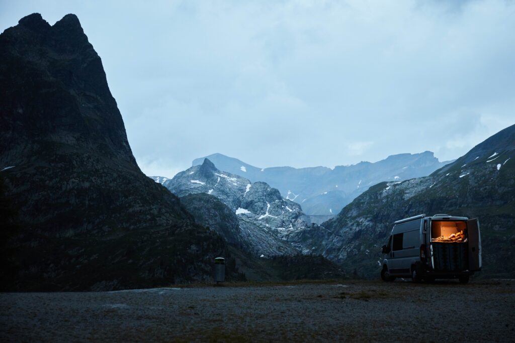 A campervan is pictured at dusk in a bleak, snowy, high-mountain setting, snow caught in the cracks of rocks. The back doors of the camper are thrown open with a soft orange light lighting up the cosy cabin, with a couple snuggling inside
