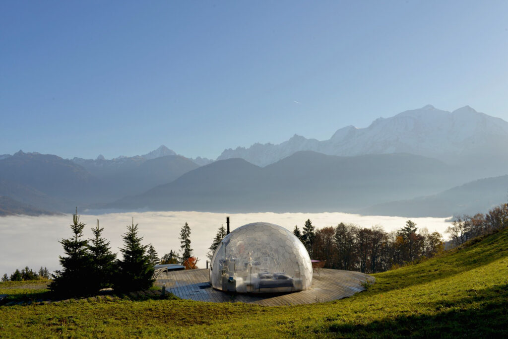 A transparent dome has a bed inside, situated on a high mountain plateau with stellar views across to jagged peaks