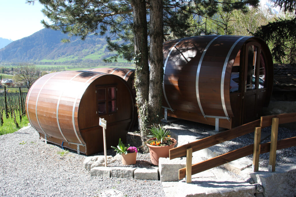 three wooden barrel shaped cabins are in a green, summery alpine setting