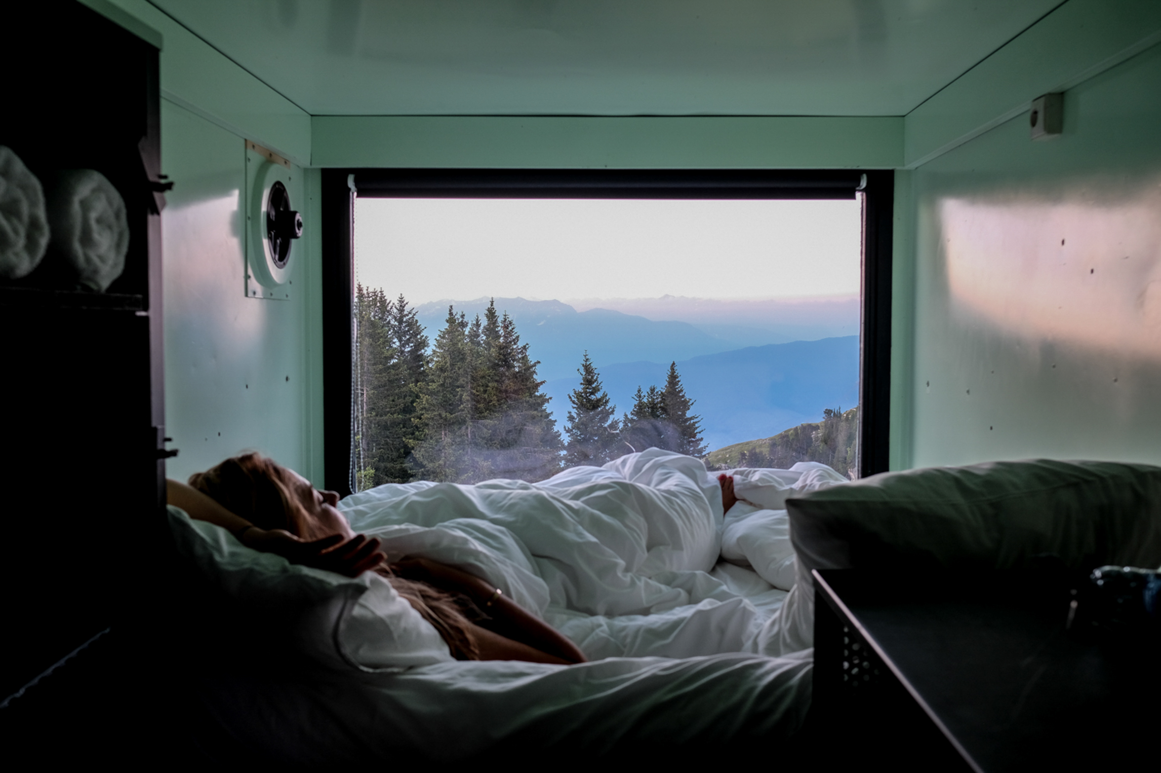 A woman lies in bed looking through the window-wall of the tiny container box she's sleeping (or woken up) in, looking over to the silhouettes of mountains with trees in the foreground