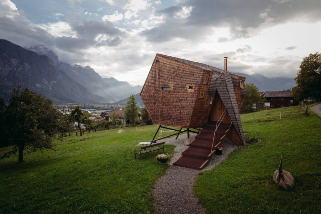 An architectural, wooden, unusually shaped cabin sits on stilts in an alpine setting, the outside tiny wooden tiles