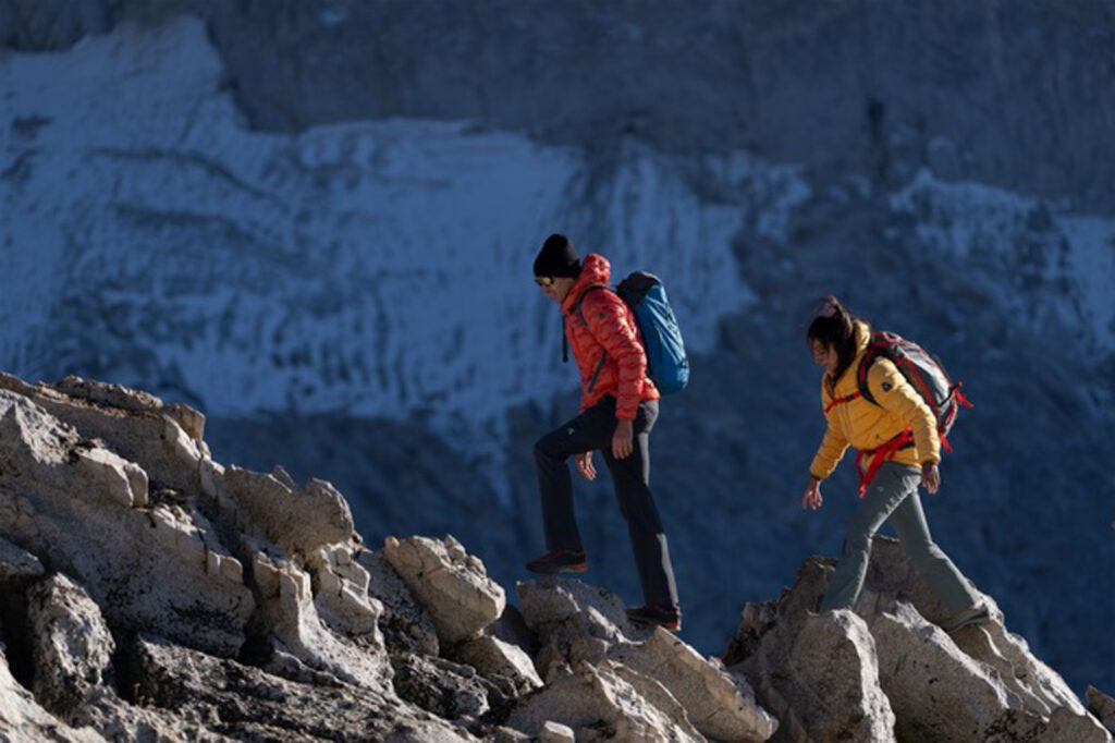 Two walkers wearing puffa jackets and backpacks walk up big rocks, a snow-dusted rock face blurred behind them