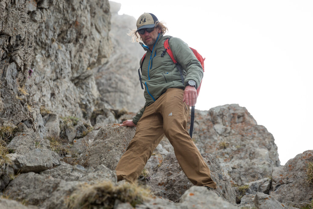 Dressed in brown pants and a sage green jacket with blue zipper, a man wearing a cap, sunnies and a red backpack walks a rocky high mountain route