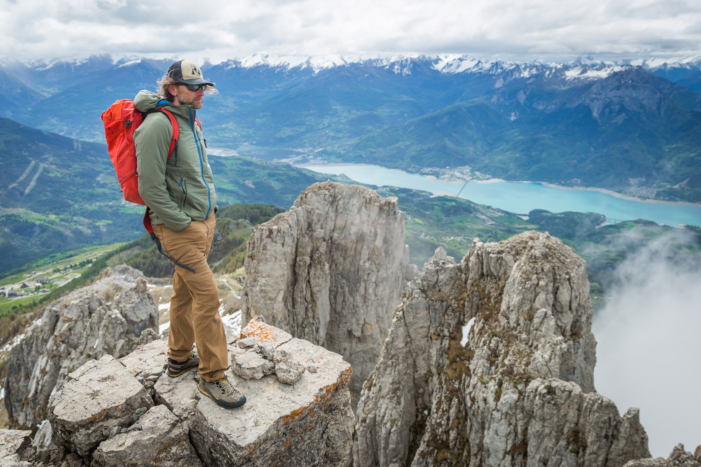 Dressed in brown pants and a sage green jacket with blue zipper, a man wearing a cap, sunnies and a red backpack stands atop a mountain on a small rock platform looking over the view, the green valley far below with a long blue lake down the middle