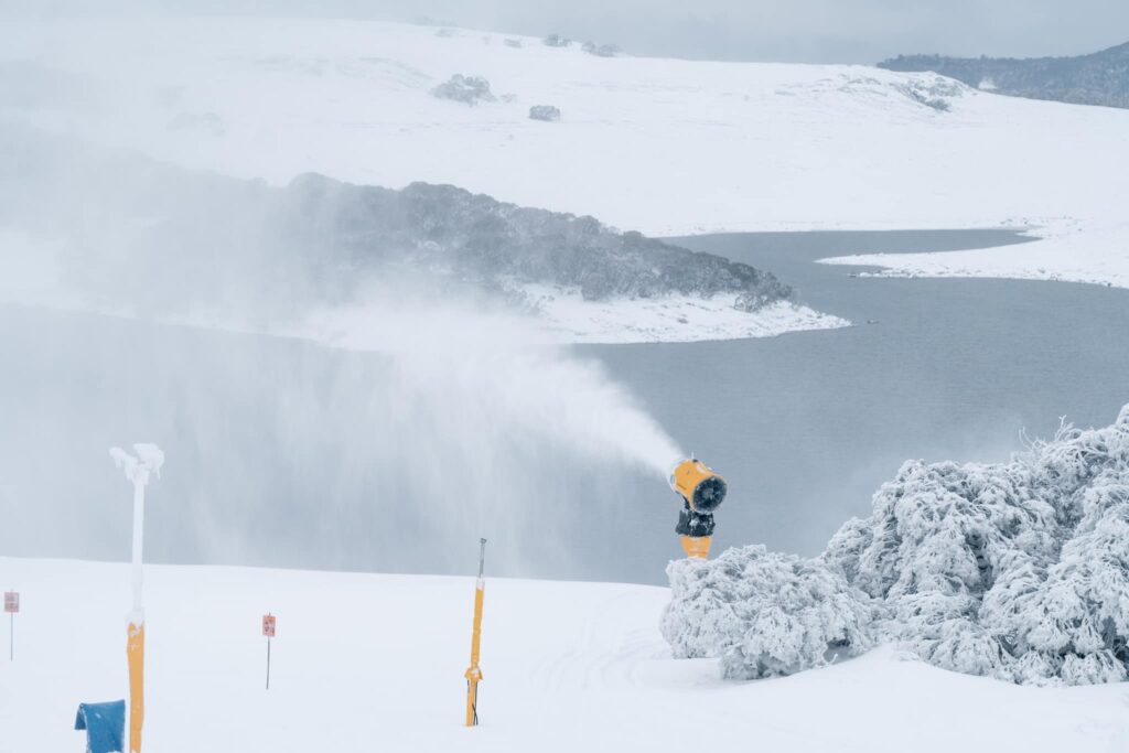 A yellow snow machine pumps out snow over a powder covered hill, a river running below
