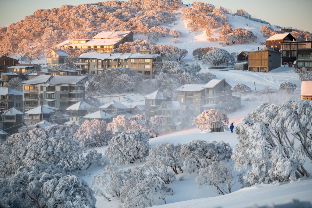 A ski village is shot (at first light) with bushy trees covered in snow and roofs dusted with powdery snow. A low sun is reflecting off the houses' windows